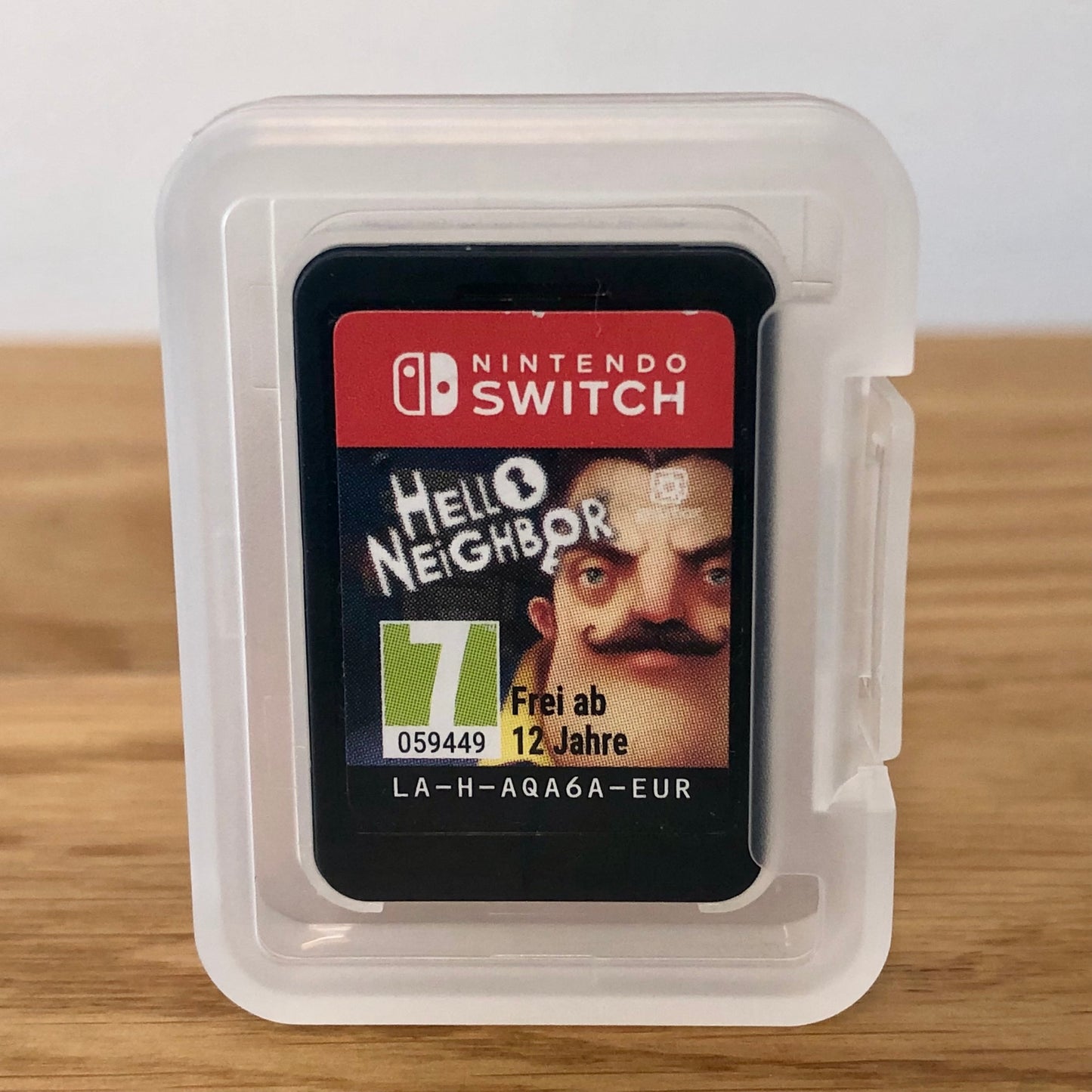 Cartridge of Hello Neighbor for Nintendo Switch, the cartridge is inside a cartridge size transparent protective case, through which you can see the front of the cartridge.