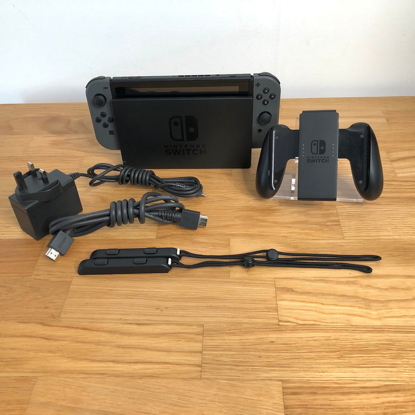 Grey Nintendo Switch console with charger, HDMI cable, Joy-Con grip and wrist straps laid out on table.