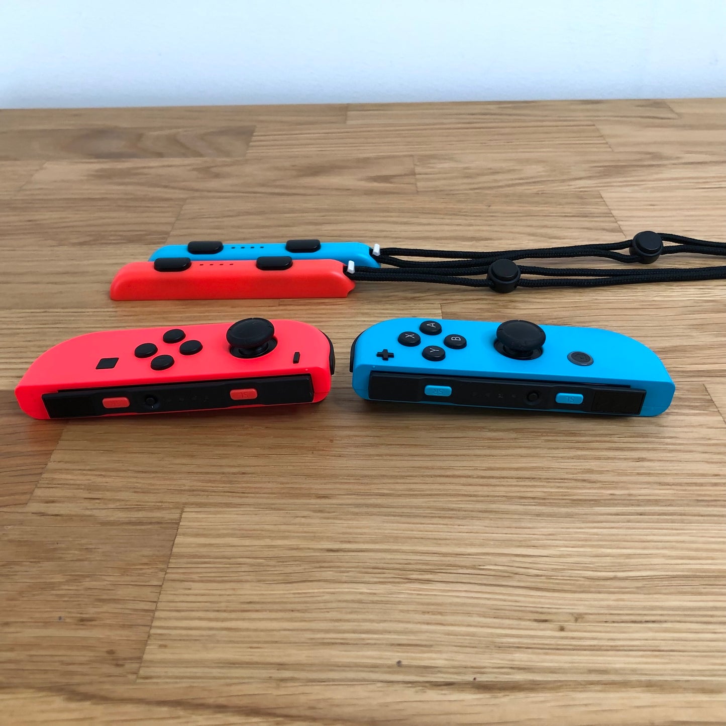 Side view of neon red and blue Nintendo Switch Joy-Con controllers lying flat with wrist straps behind.