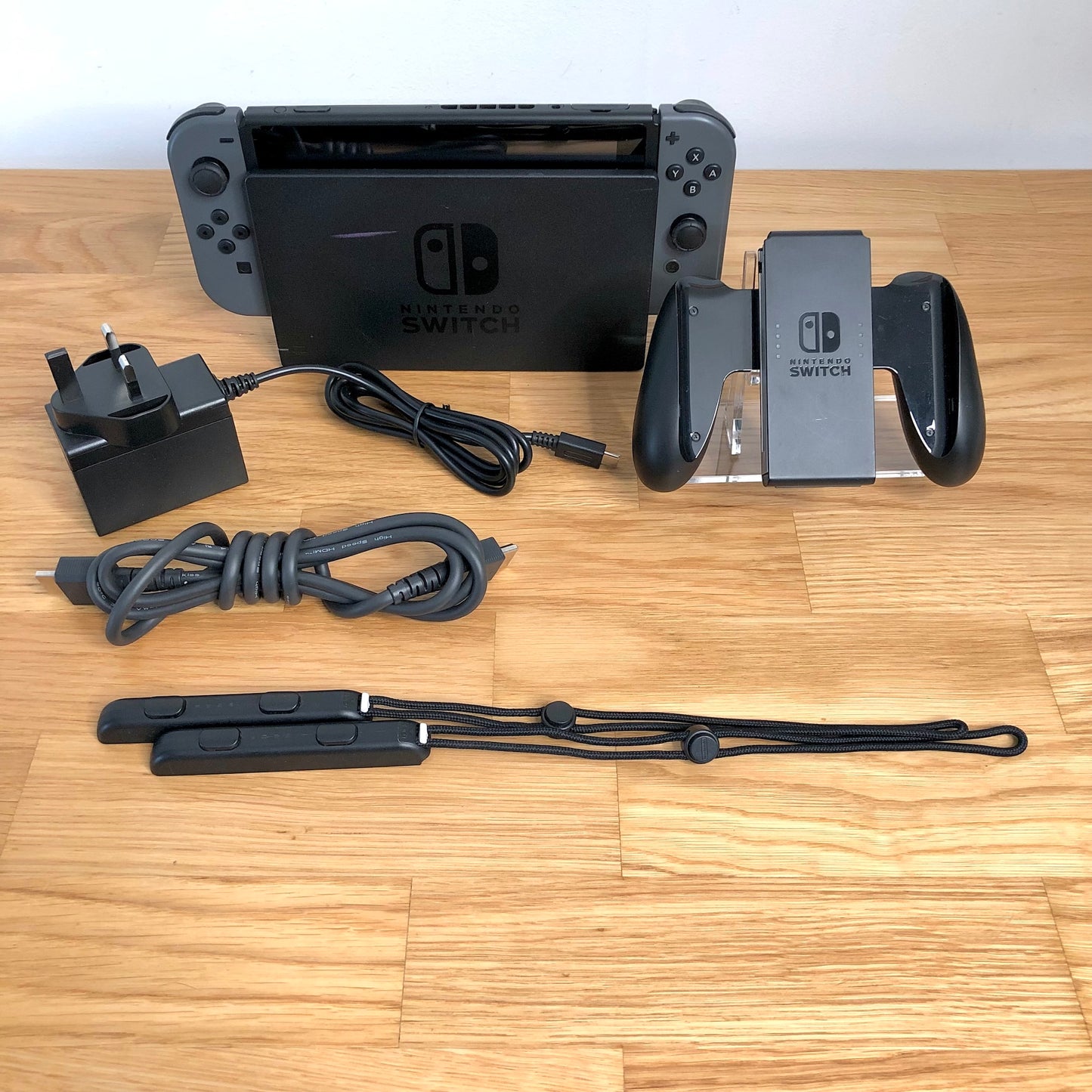 Grey Nintendo Switch console with charger, HDMI cable, Joy-Con grip and wrist straps laid out on table.