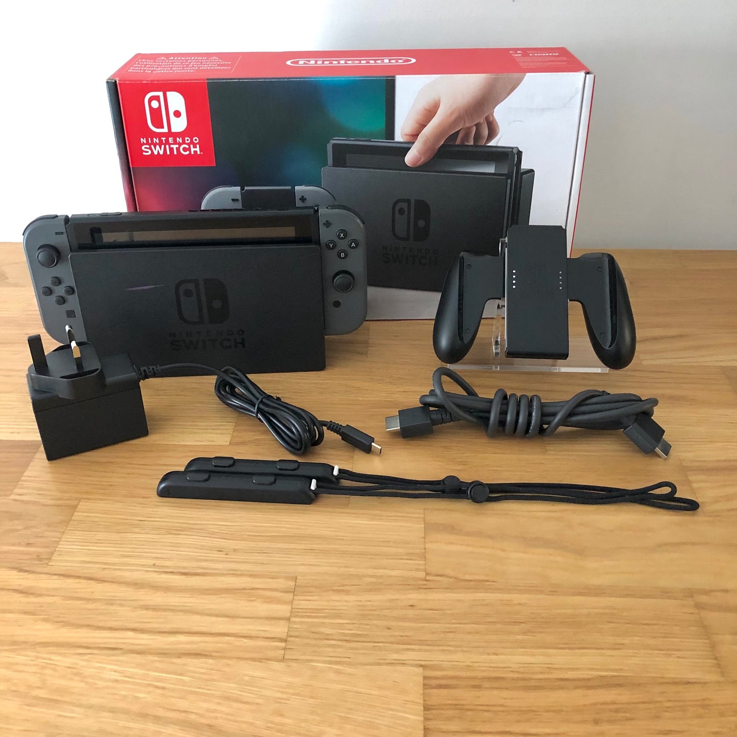 Grey Nintendo Switch console with original box, Joy-Con grip, charger, HDMI cable and wrist straps shown.