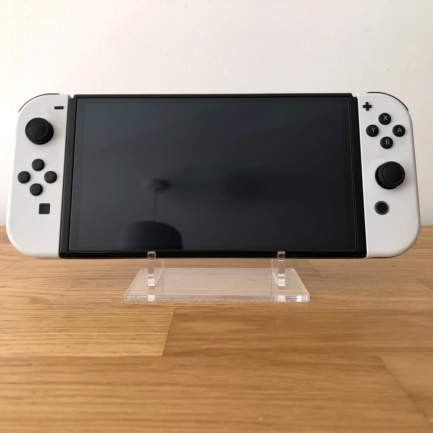 Front view of Nintendo Switch OLED console with white Joy-Cons attached, screen off, on clear display stand.