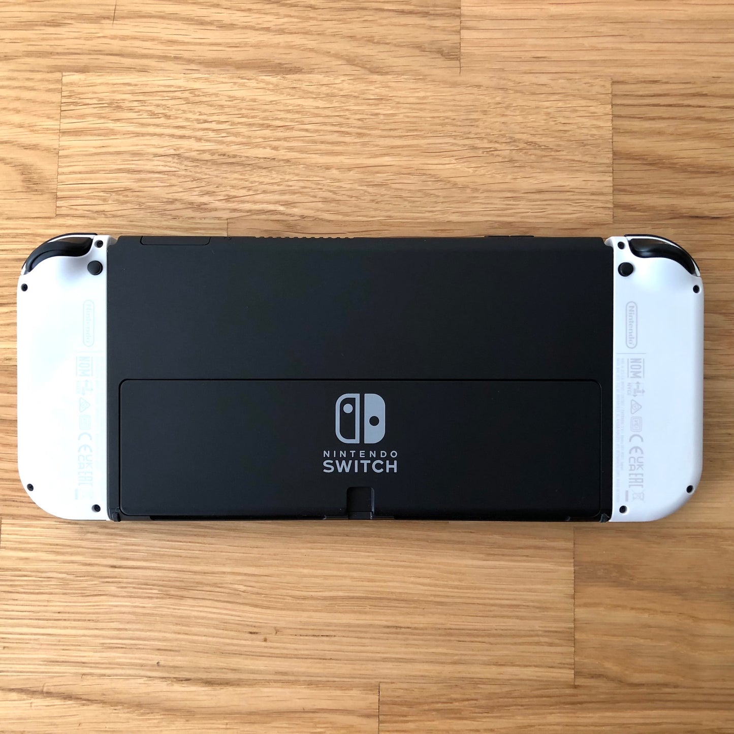 Back of Nintendo Switch OLED console with white Joy-Cons shown flat on table, logo and kickstand visible.