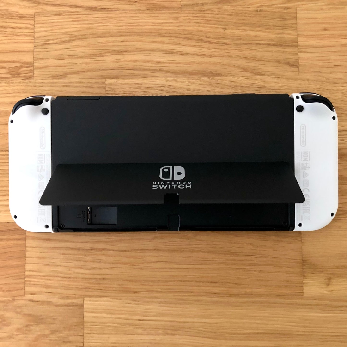 Back view of Nintendo Switch OLED with kickstand lifted, showing microSD slot, USB-C port and controls.