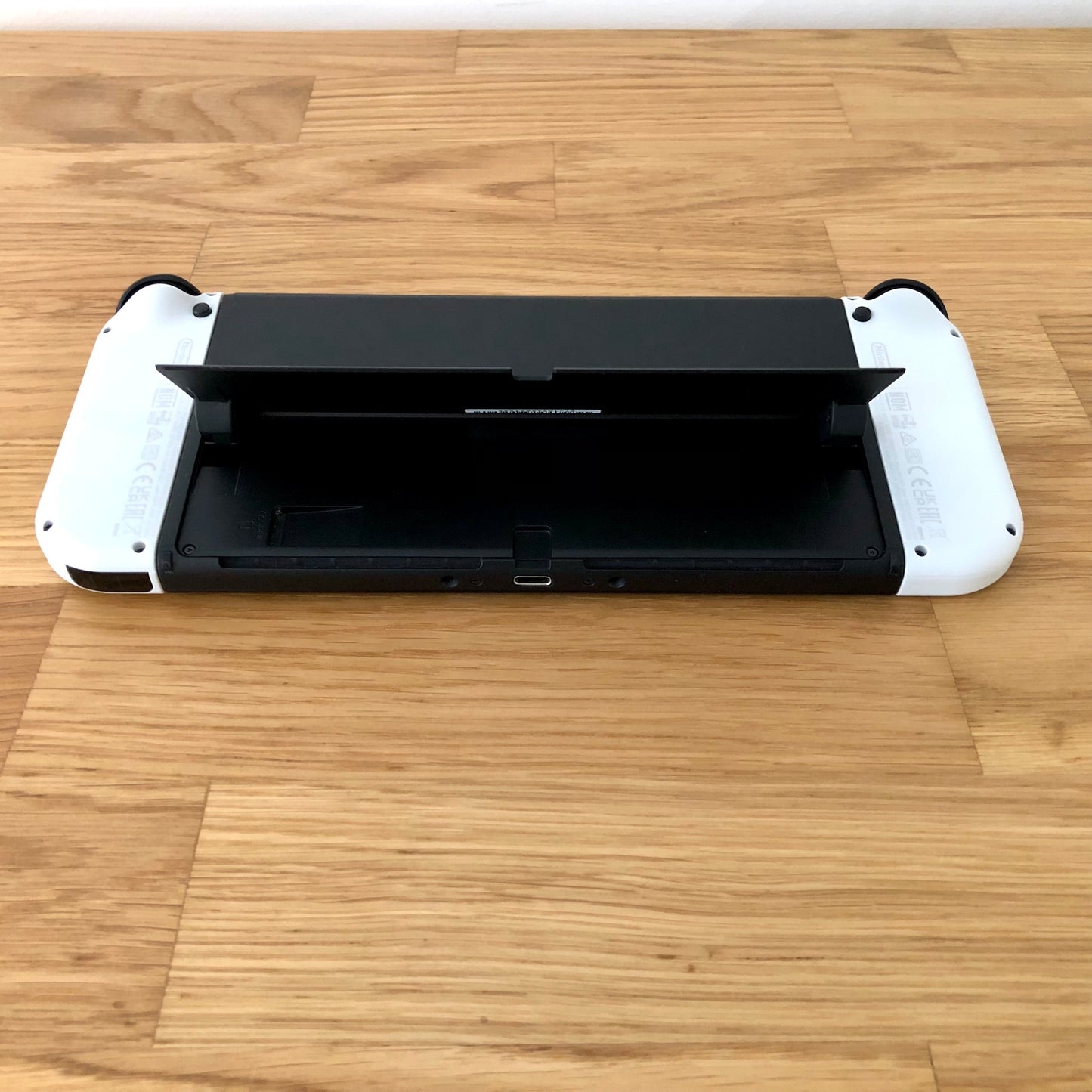 Bottom rear view of Nintendo Switch OLED with kickstand lifted, showing microSD slot, USB-C port and controls.
