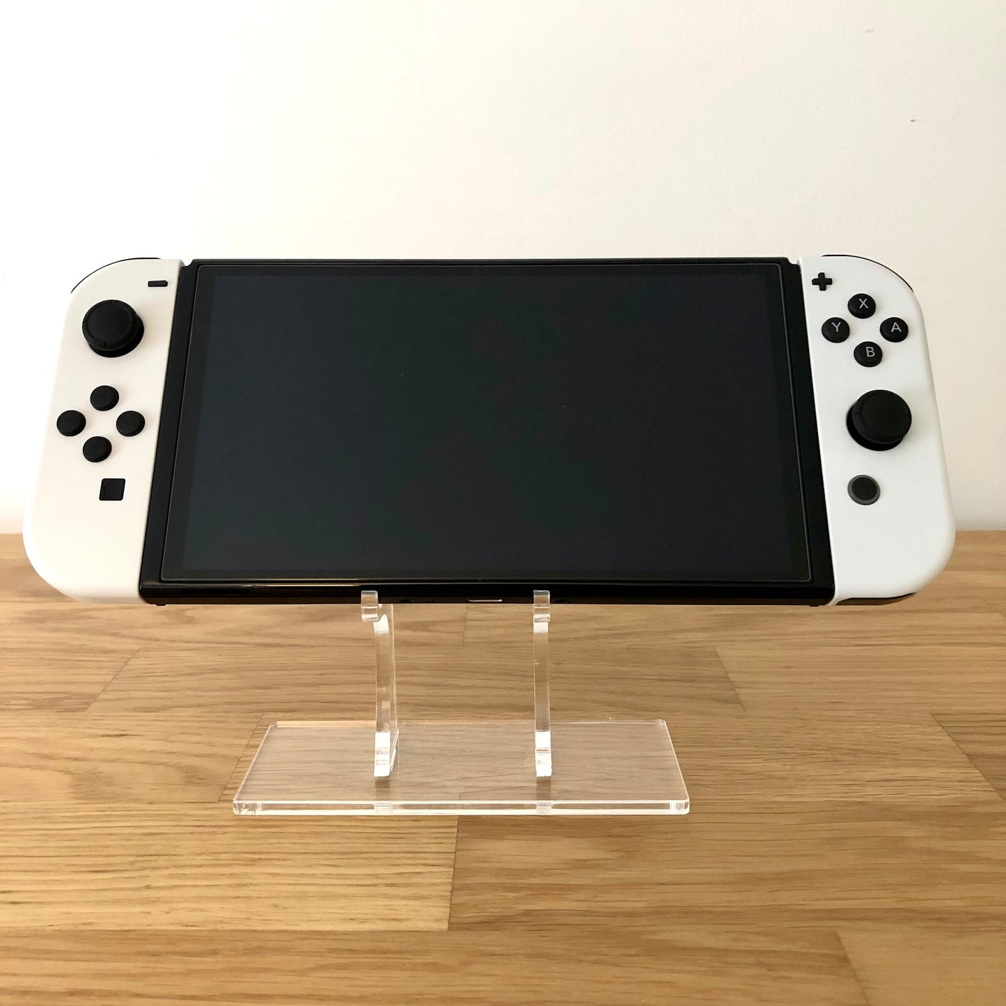 Front view of Nintendo Switch OLED console with white Joy-Cons attached, screen off, on clear display stand.