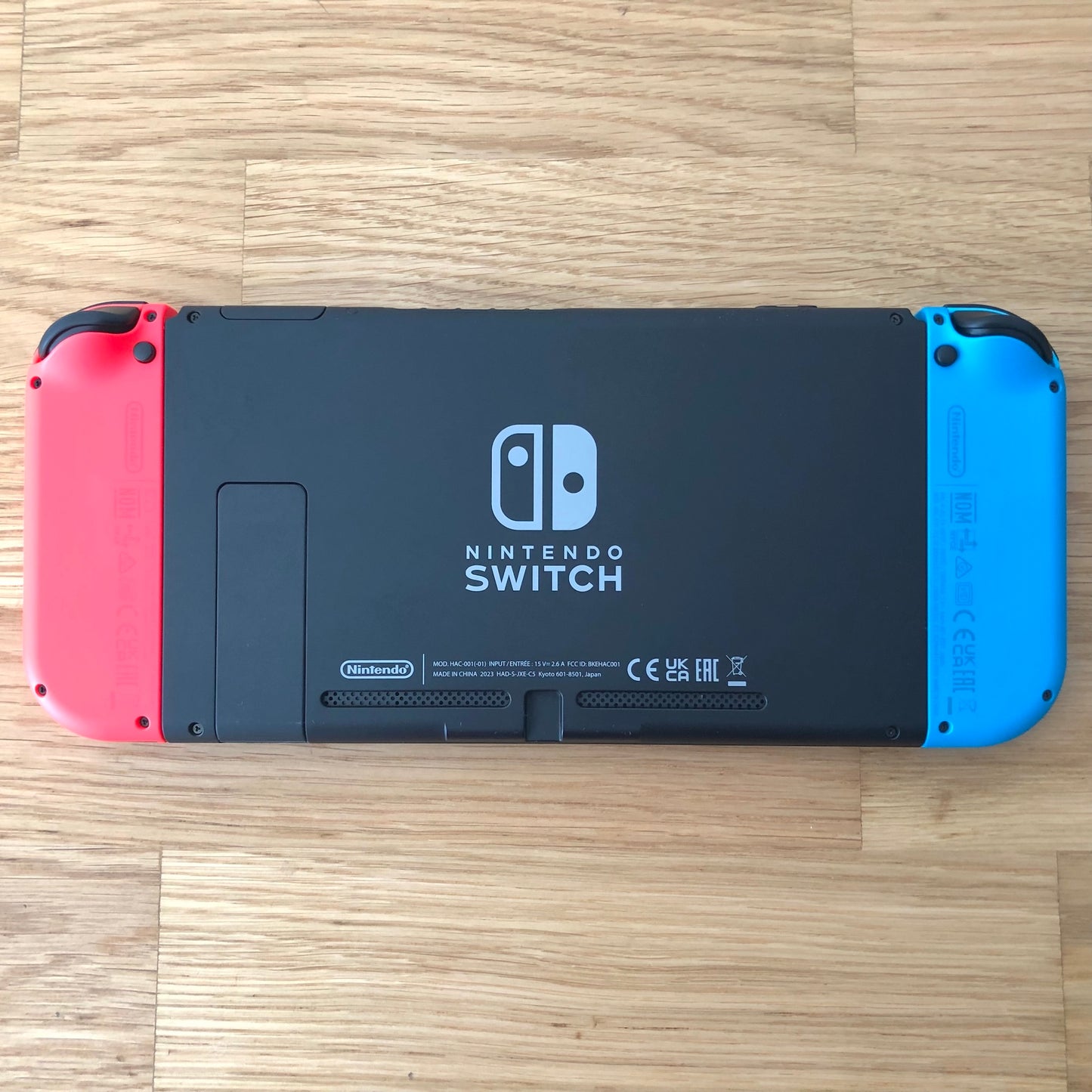 Back of Nintendo Switch console with red and blue Joy-Cons attached, showing logo, kickstand and trigger buttons. Console laid flat.
