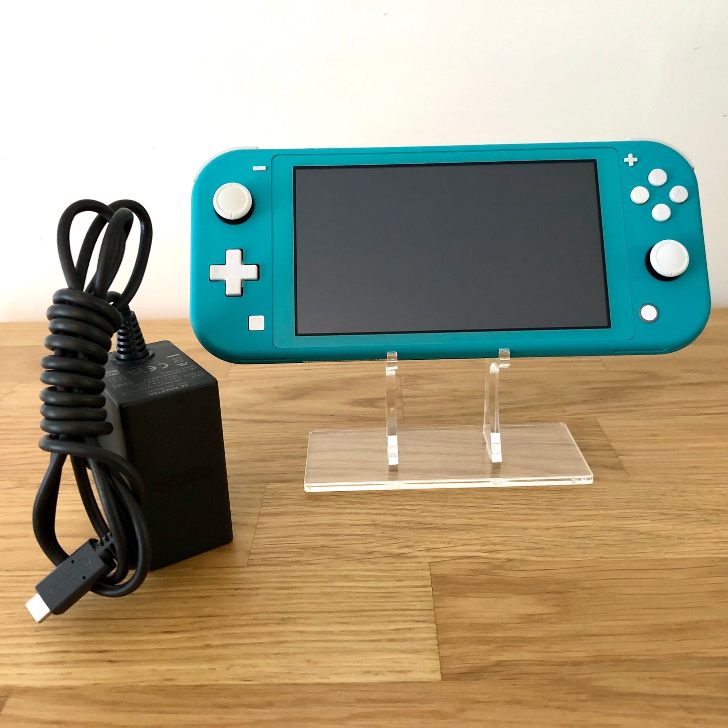 Front of Nintendo Switch Lite in Turquoise, displayed on a stand with official charger placed beside it on a wooden surface.