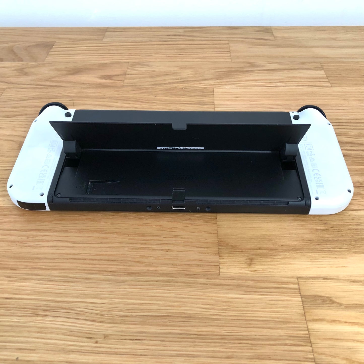 Bottom rear view of Nintendo Switch OLED with kickstand lifted, showing microSD slot, USB-C port and controls.