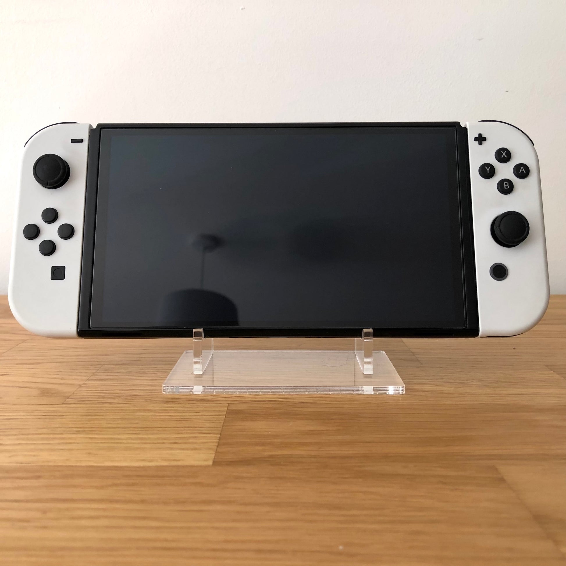 Front view of Nintendo Switch OLED console with white Joy-Cons attached, screen off, on clear display stand.