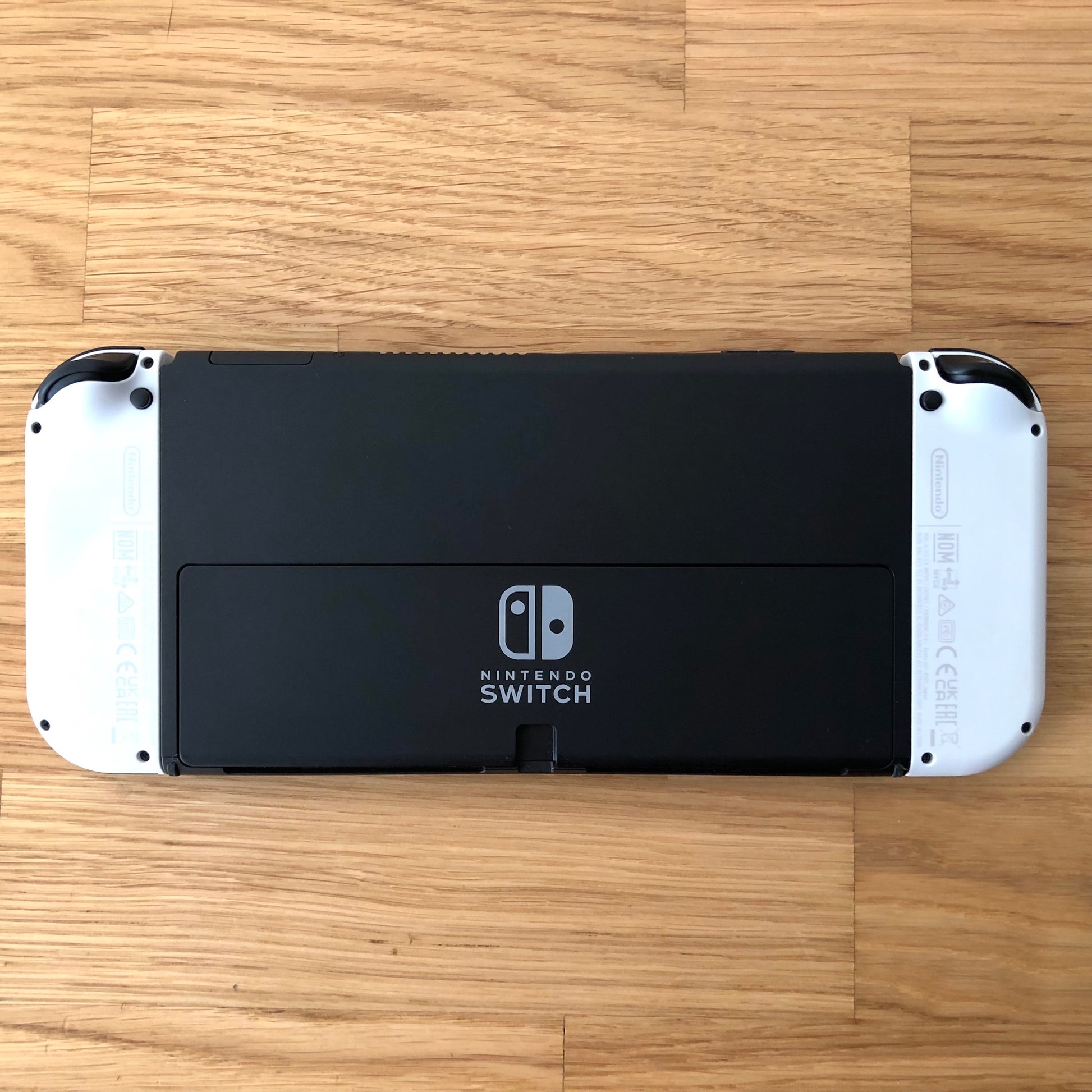Back of Nintendo Switch OLED console with white Joy-Cons shown flat on table, logo and kickstand visible.