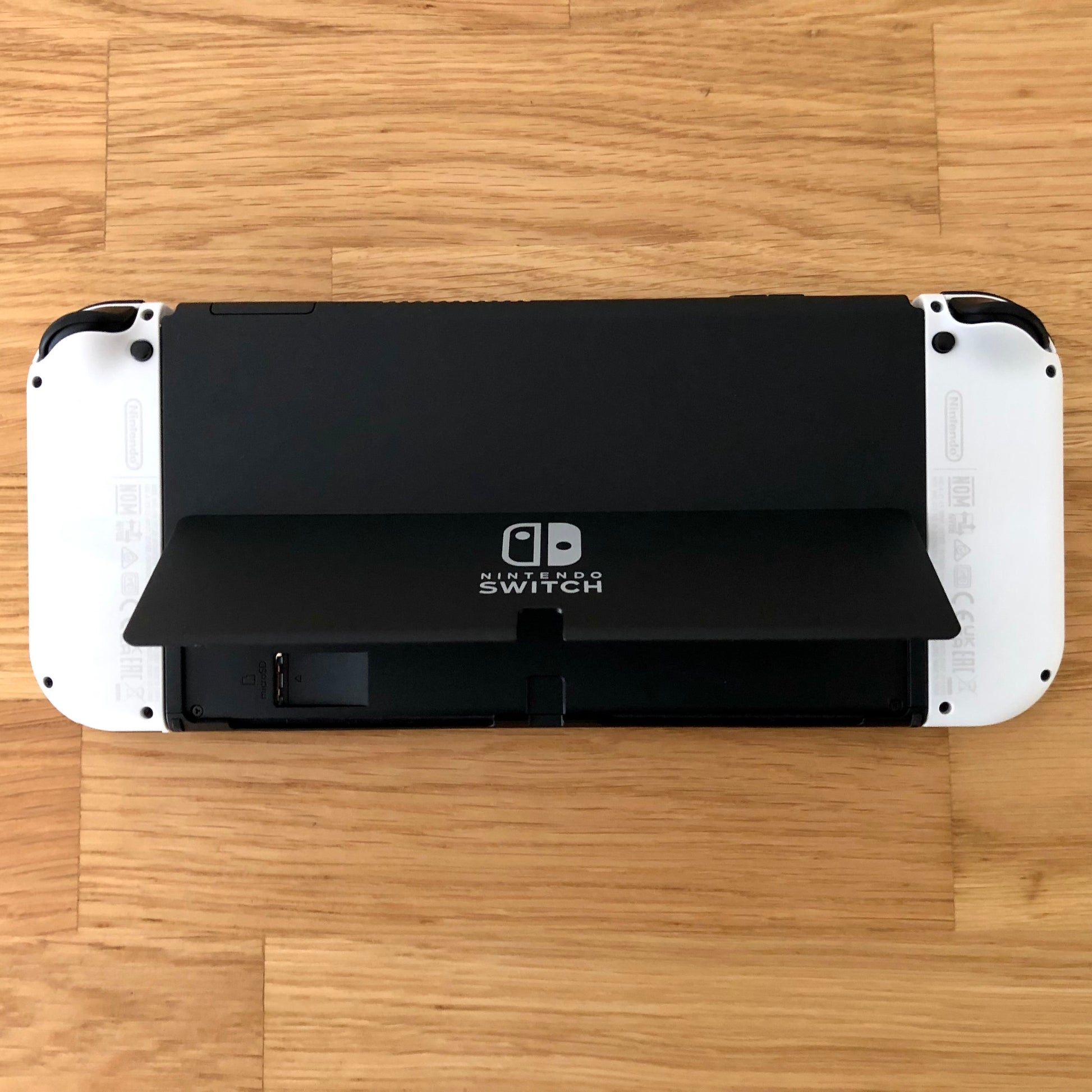 Back view of Nintendo Switch OLED with kickstand lifted, showing microSD slot, USB-C port and controls.