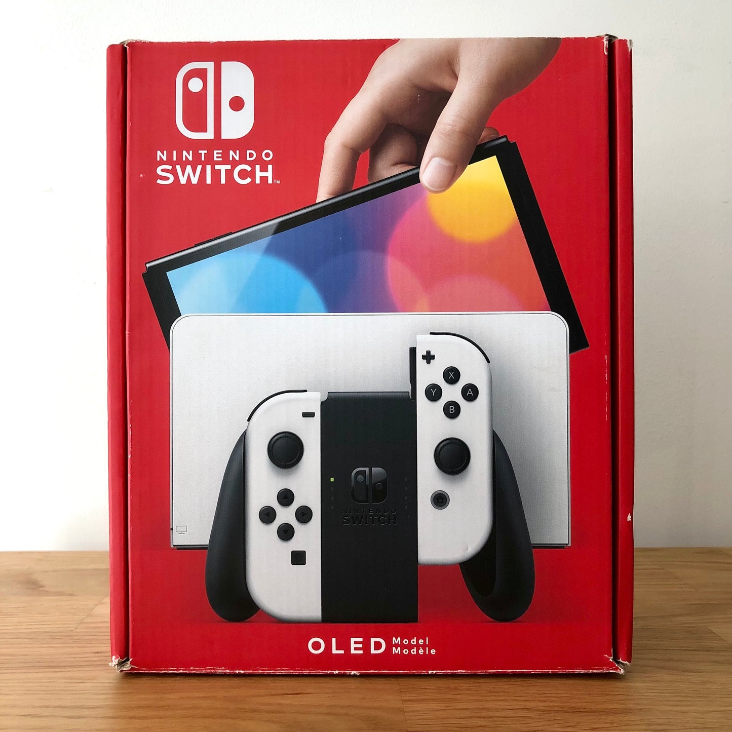 Front of Nintendo Switch OLED box showing white console and Joy-Cons, with colourful screen artwork and red background.