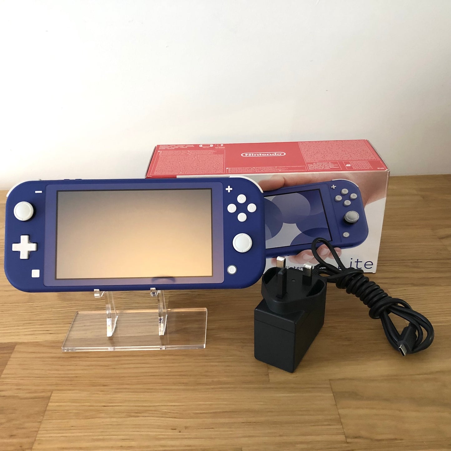 Blue Nintendo Switch Lite console with charger and original box, displayed upright on a clear stand.