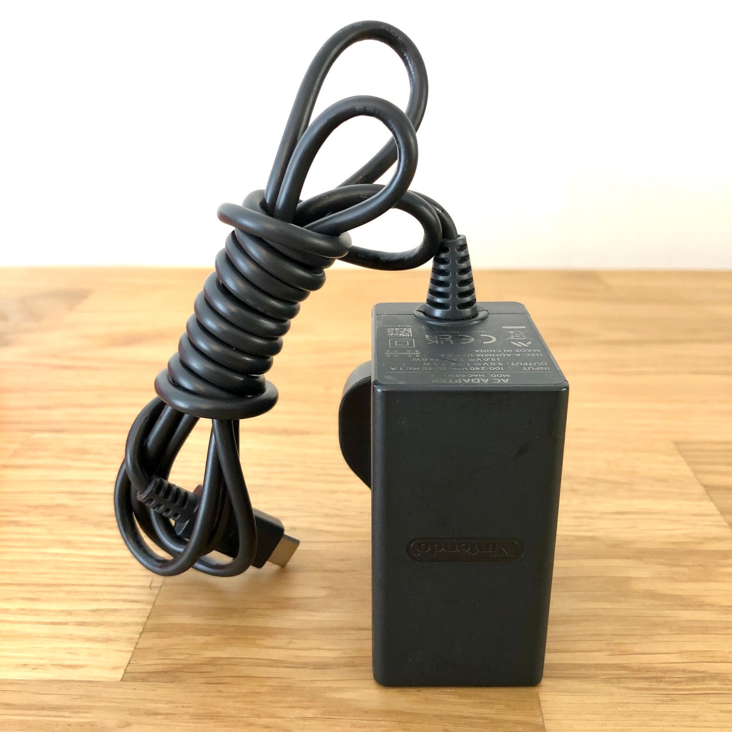 Official Nintendo Switch AC charger with UK plug and USB-C cable, shown coiled neatly on a wooden surface.