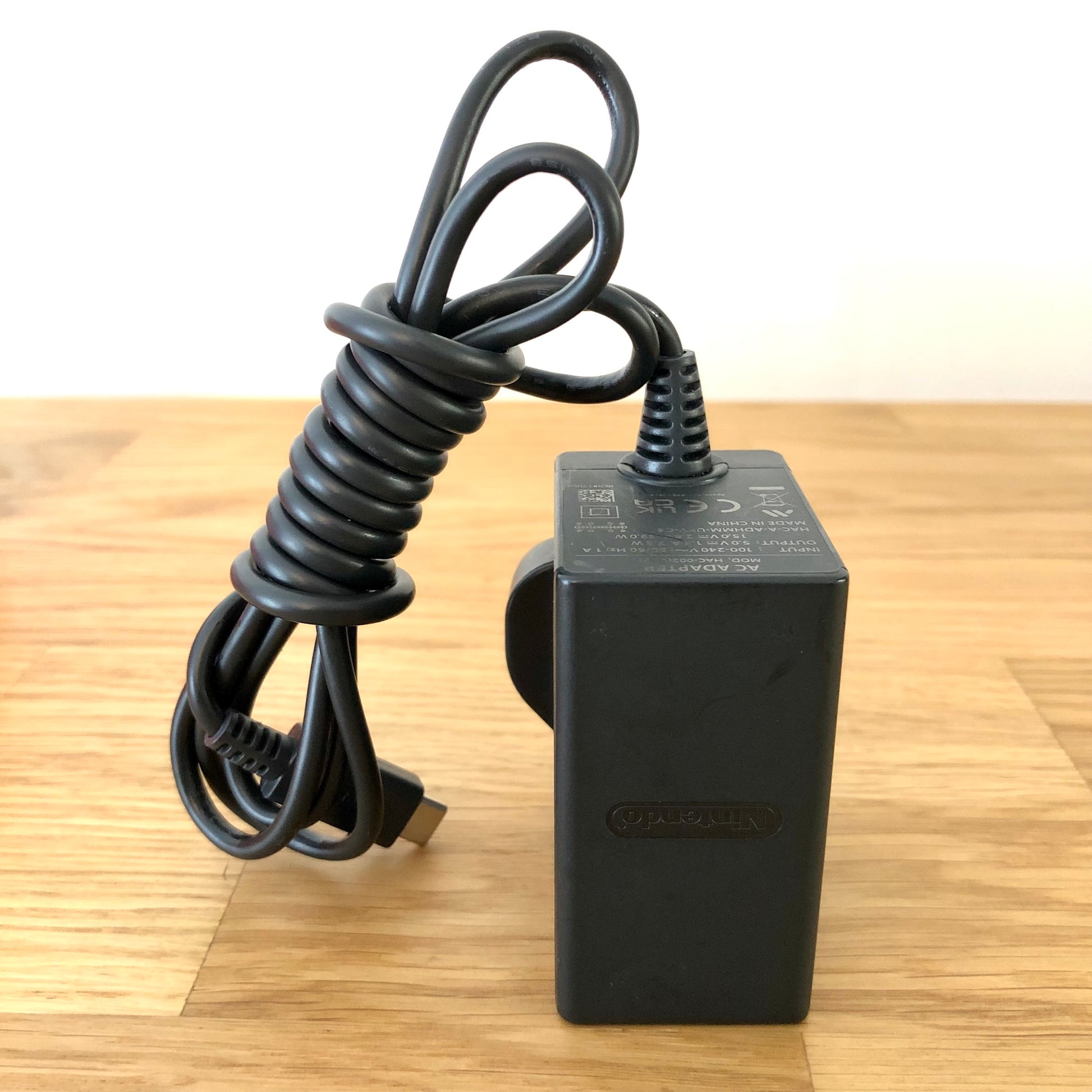 Official Nintendo Switch AC charger with UK plug and USB-C cable, shown coiled neatly on a wooden surface.